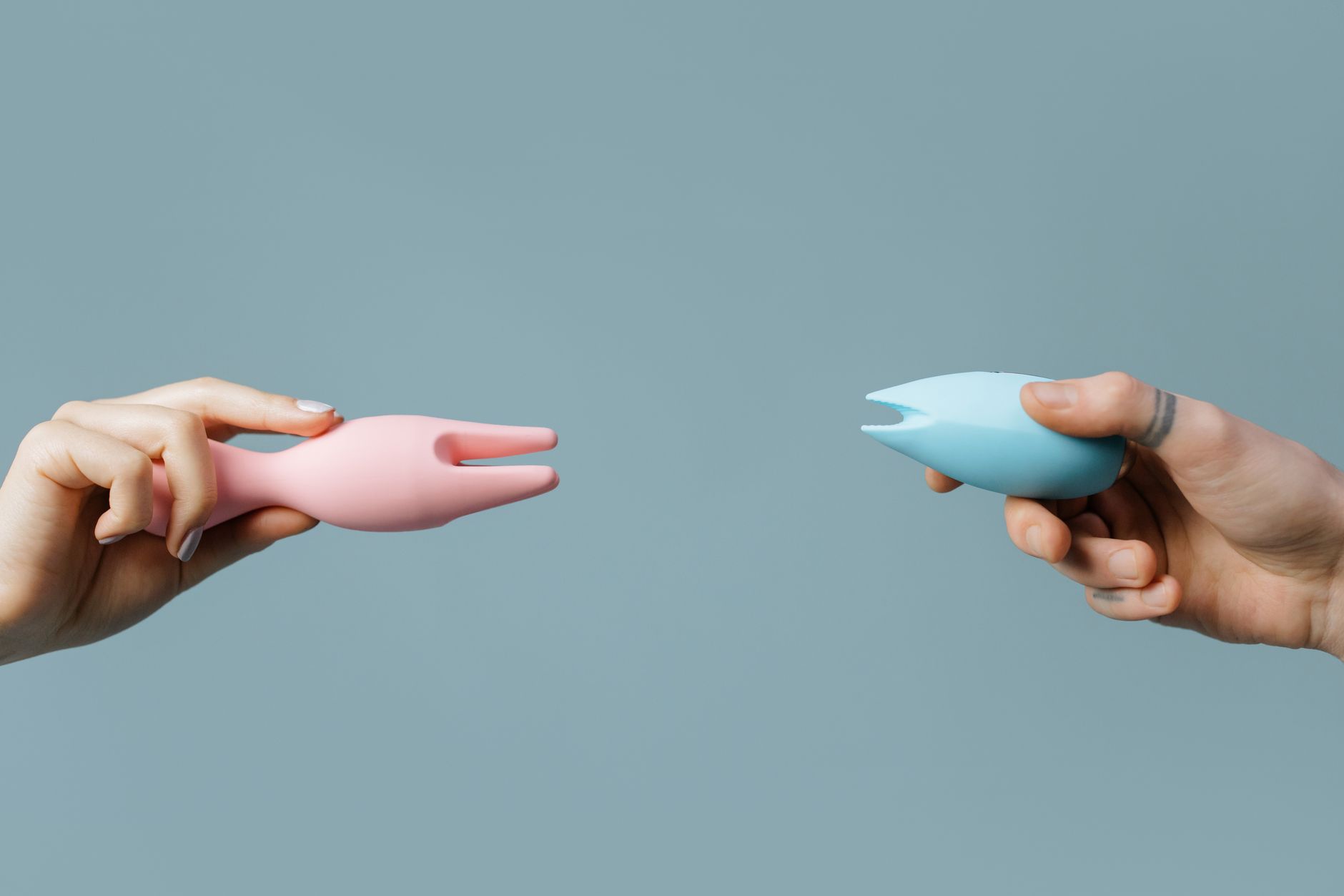 Two hands holding pink and blue lemon-shaped silicone vibrators against a soft pastel background