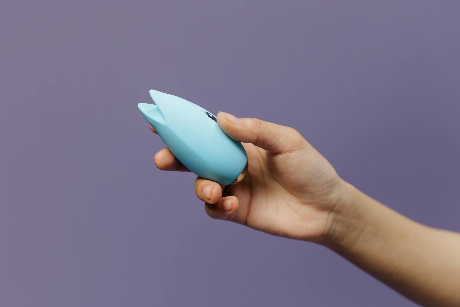 A blue silicone clitoral vibrator held in hand against a solid purple background, promoting self-love and solo pleasure.
